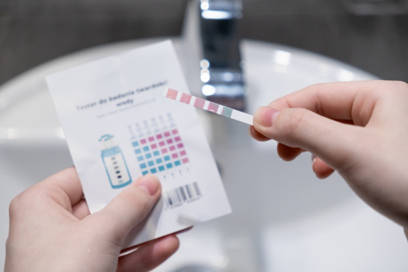 A close-up of hands holding a water test strip and color chart, comparing results to measure water hardness (Polish) for home purity and health