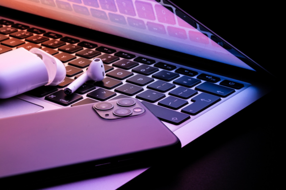 a laptop half closed in the dark with a phone and wireless earphones glowing