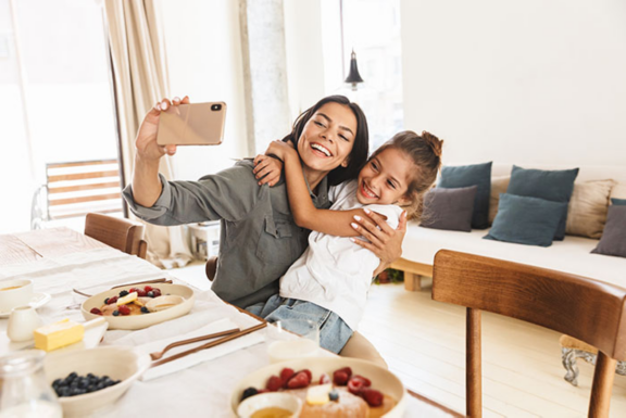 Image of smiling family mother and little daughter hugging and taking selfie photo on cellphone while having breakfast at home in morning