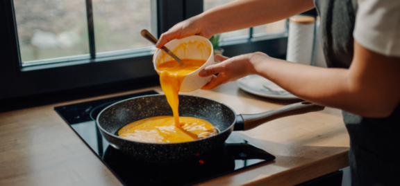 Chef pours whisked eggs from a small bowl into a frying pan on an induction stove top, preparing to cook an omelet in a modern kitchen with natural light