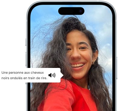 Un iPhone 15 montrant la fonctionnalité VoiceOver énonçant les informations figurant sur l’image : Une personne aux cheveux noirs ondulés en train de rire